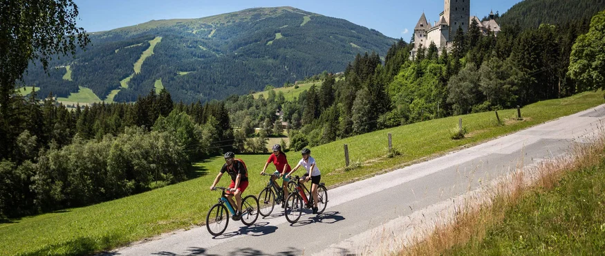 Drei Radfahrer fahren auf Landstraße mit Schloss und Bergen im Hintergrund