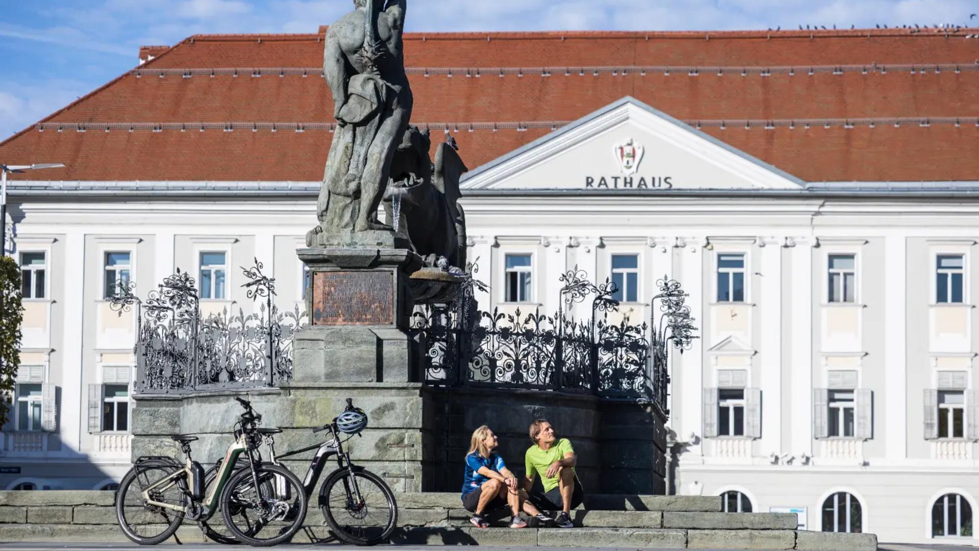 Zwei Menschen sitzen neben Fahrrädern vor einem Denkmal und Rathausgebäude