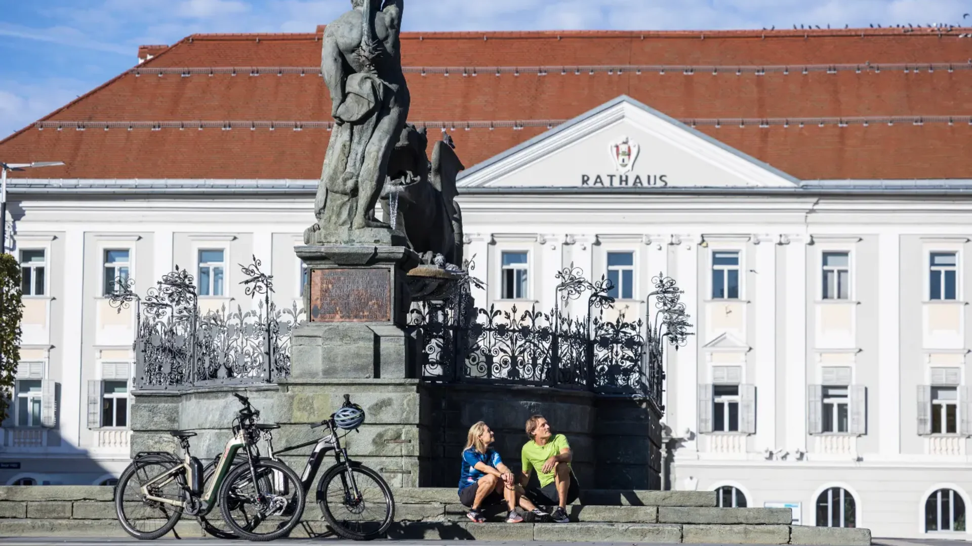 Tour-Detail © Lukas Krische Zwei Menschen sitzen neben Fahrrädern vor einem Denkmal und Rathausgebäude