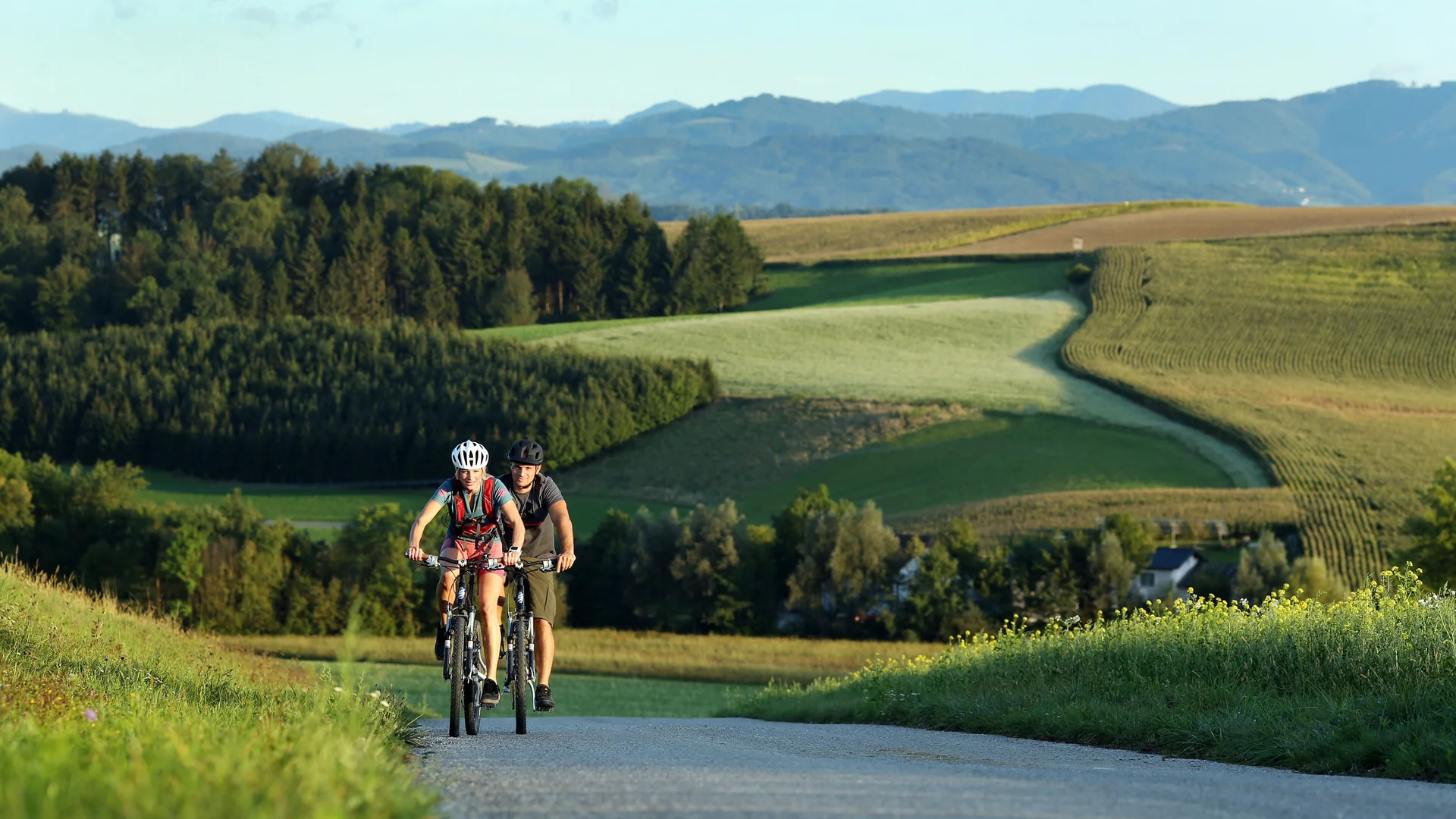 Zwei Radfahrer fahren auf Landstraße in hügeliger Landschaft bei Sonnenschein