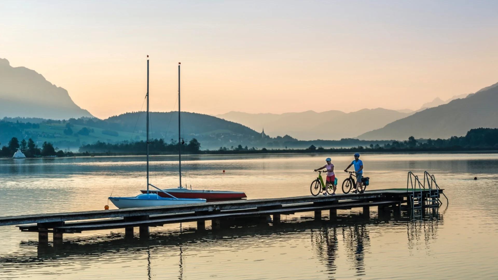 Twee fietsers op steiger bij meer met zeilboten en bergen op de achtergrond