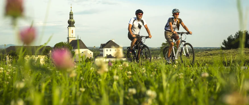 Anthering: Radurlaub zwischen Moor & Mozartstadt © Markus Greber Zwei Radfahrer fahren im Grünen vor Dorfkirche bei klarem Himmel