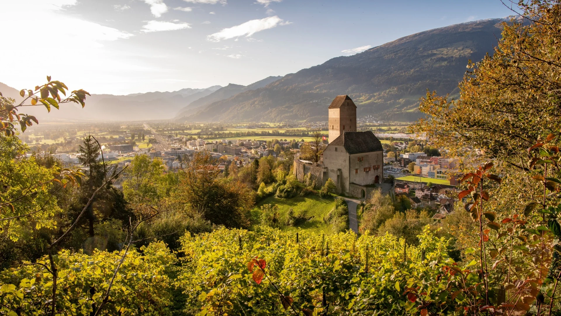 Blick auf eine Burg auf einem Hügel mit Stadt und Bergen im Hintergrund bei Sonnenlicht