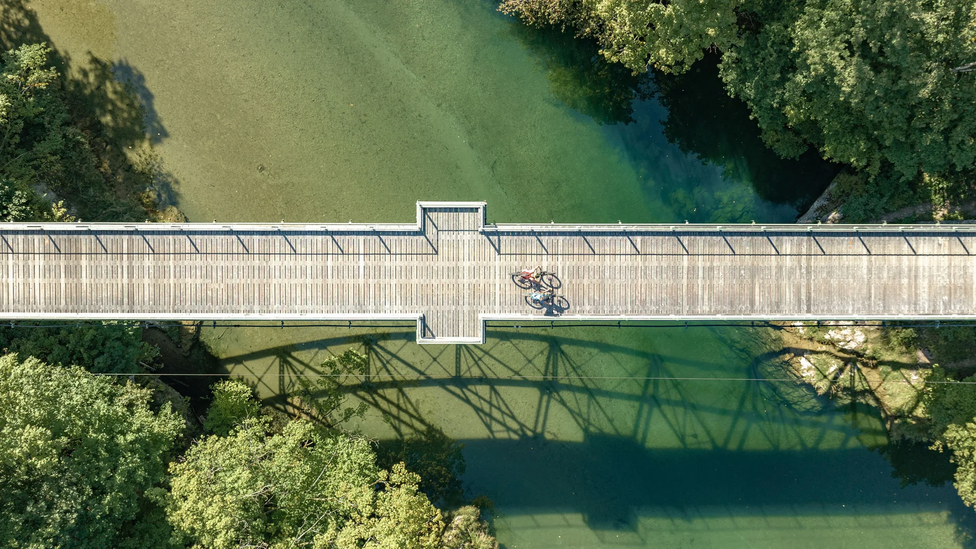 Luftaufnahme einer Holzbrücke über grünem Wasser mit zwei Radfahrern