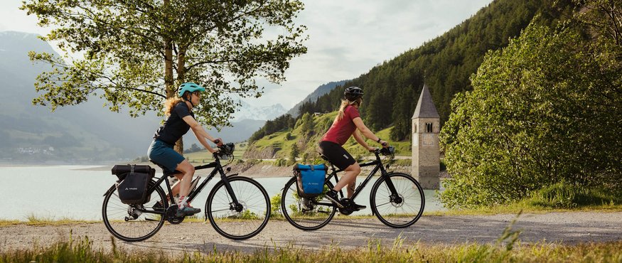 Two cyclists ride by a lake with mountain view and church tower