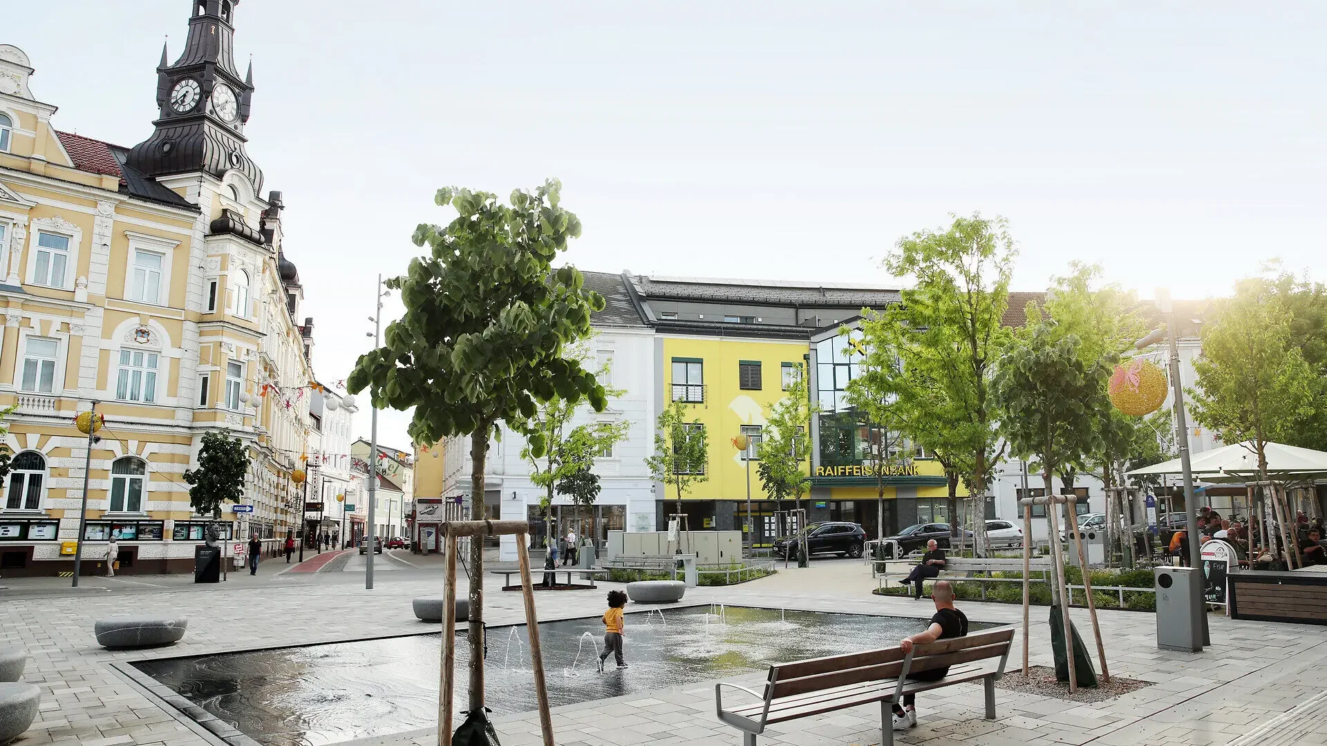 Stadtplatz mit Brunnen, historischem Uhrturm, Bäumen und Sitzbänken