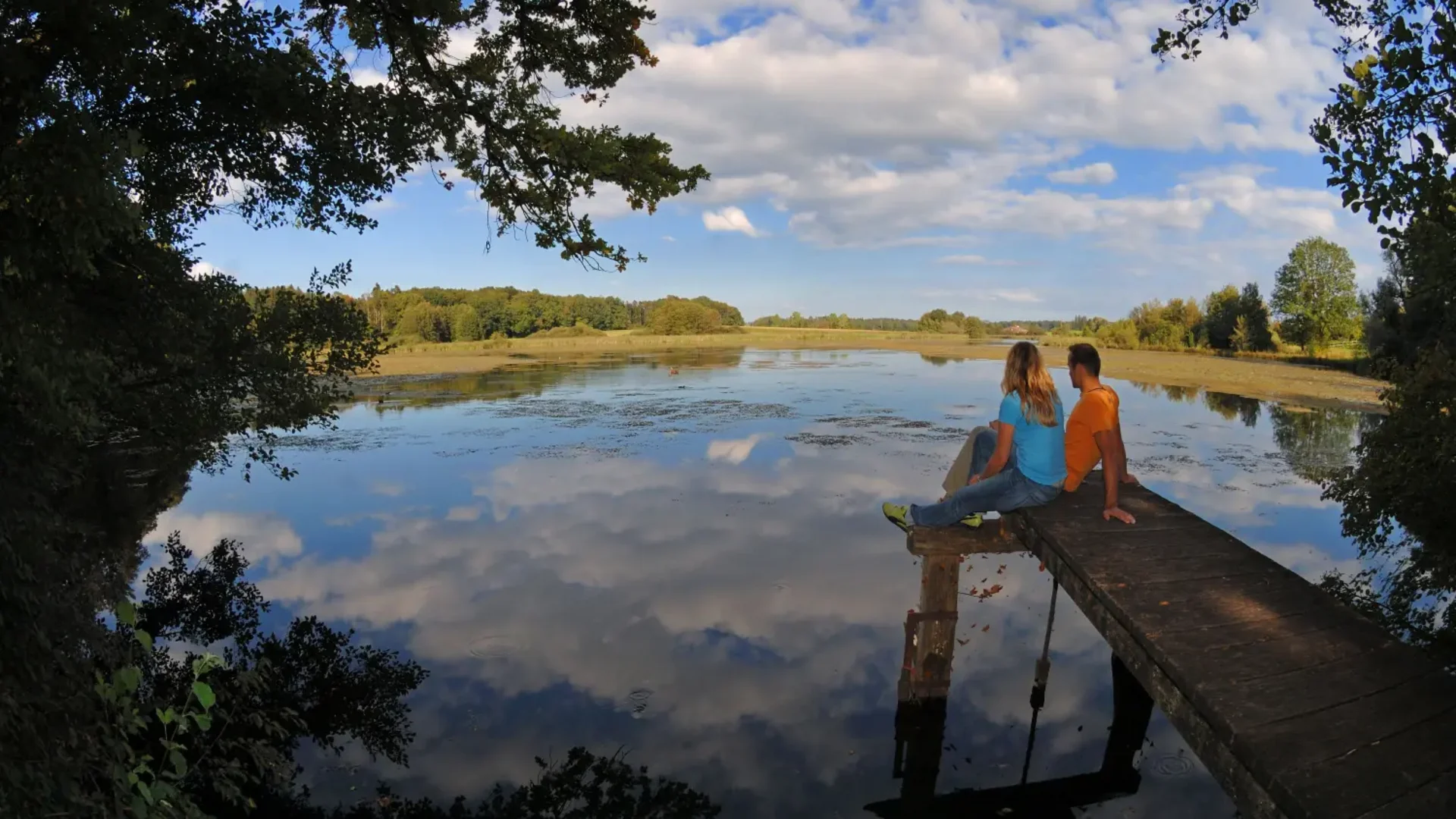 Twee personen zitten op een steiger bij een kalm meer onder een bewolkte lucht