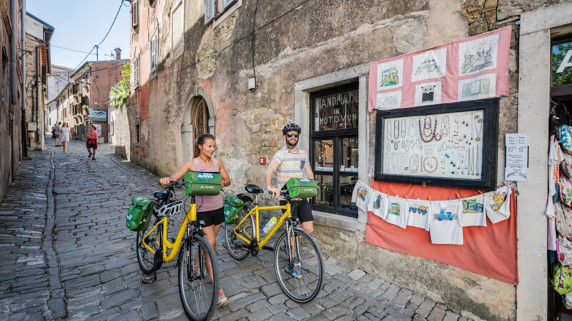 Two cyclists with yellow bikes stop to look at jewelry displayed outside a shop on cobblestone street
