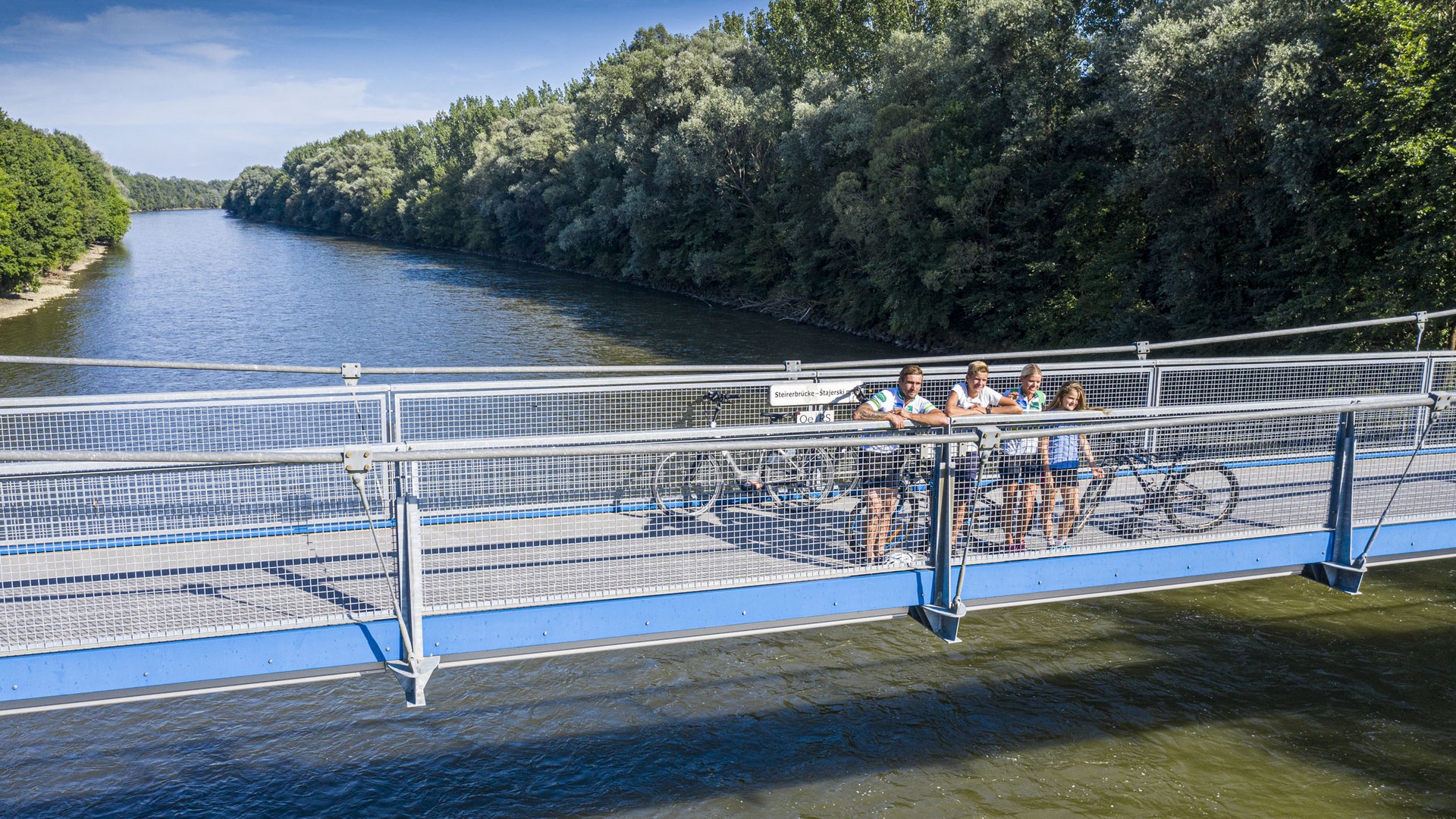 Cyclists standing on blue bridge over river with trees on riverbank