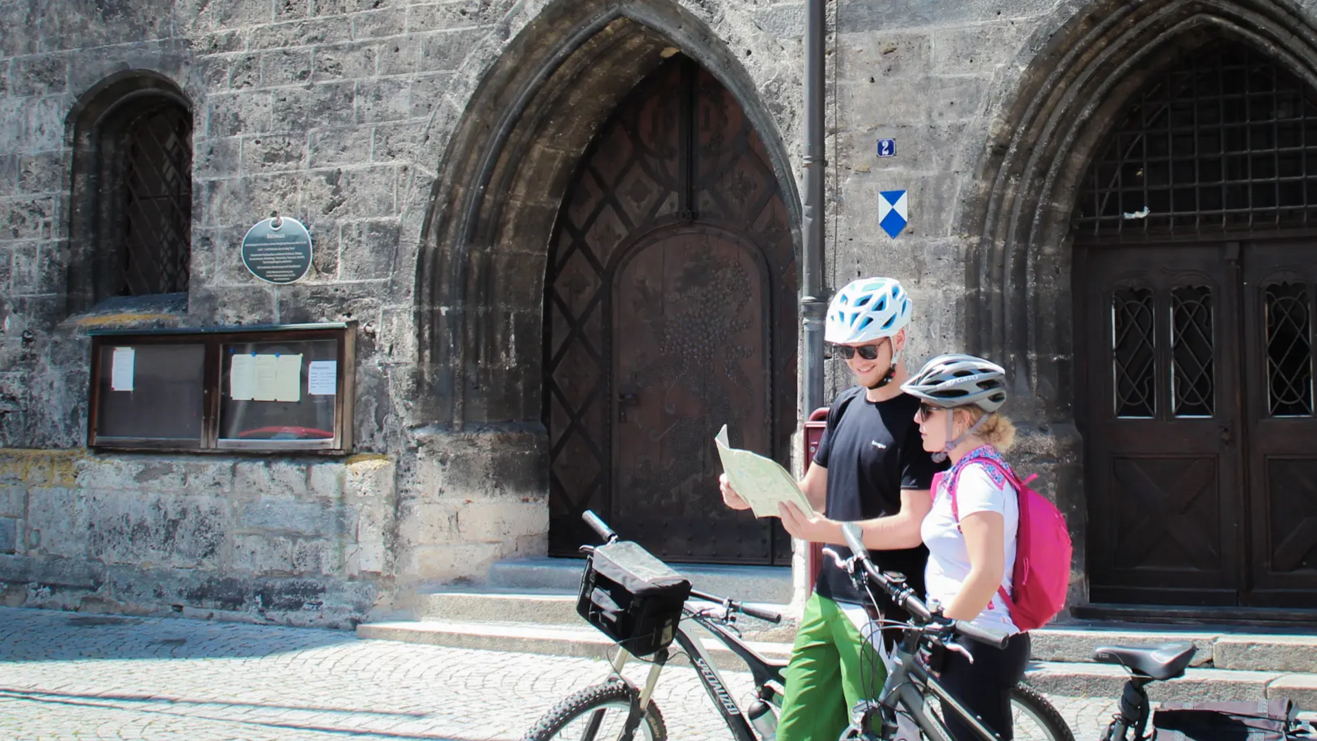 Two cyclists with helmets reading map in front of old stone portal