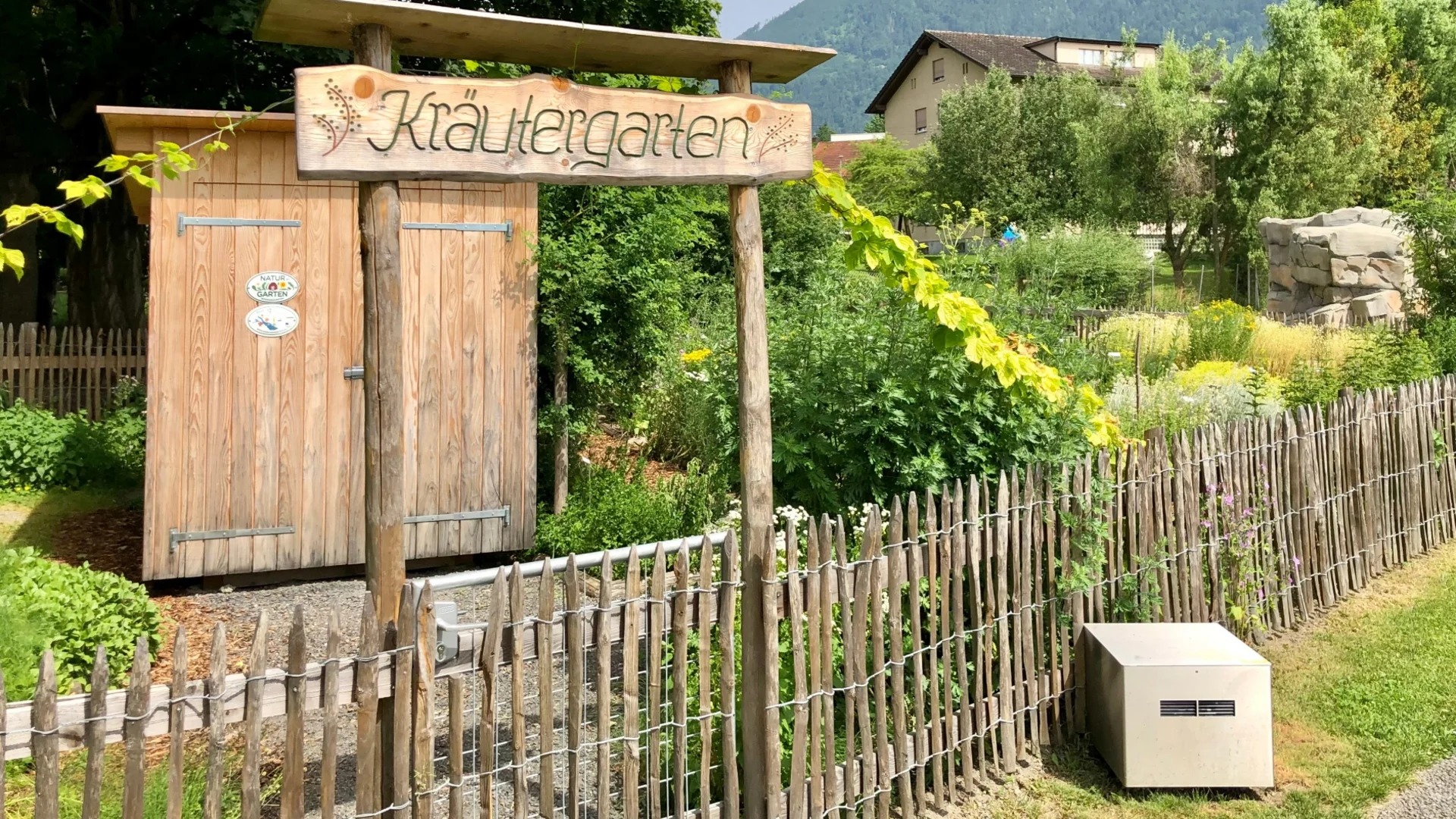 Hölzernes Schild Kräutergarten vor Zaun und Garten mit Bergen im Hintergrund
