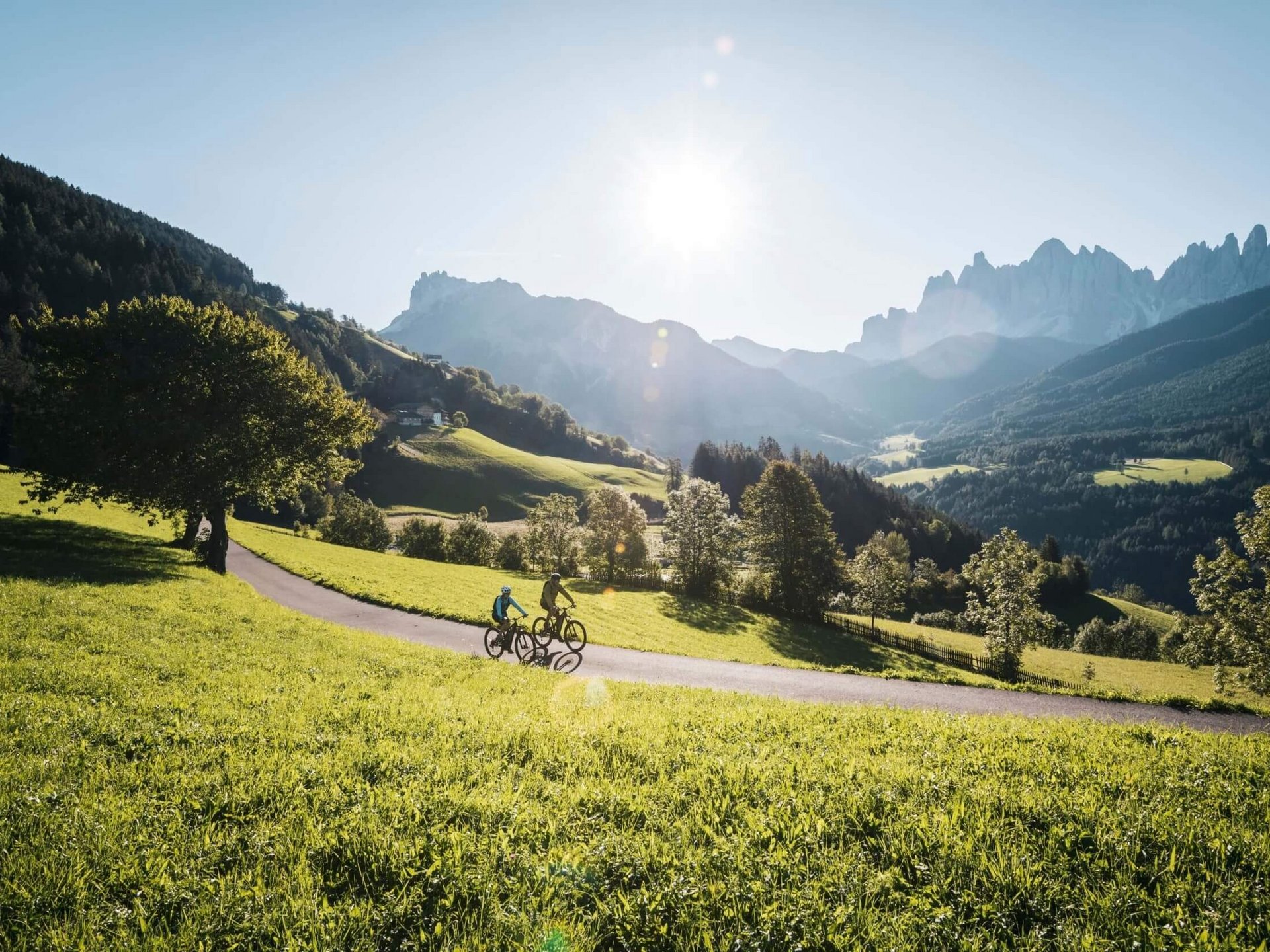 Two cyclists riding on a path through green alpine landscape under sunny sky
