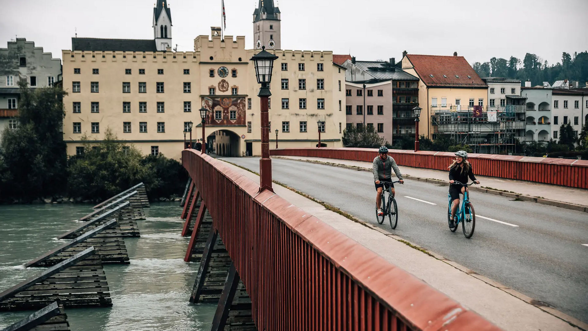 Two cyclists on red bridge by historic buildings and river