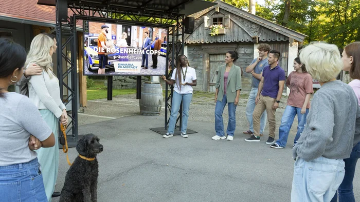 Filmreife Radelpause © Bavaria Filmstadt Eine Gruppe hört einer Präsentation im Freien zu; daneben sitzt ein Hund