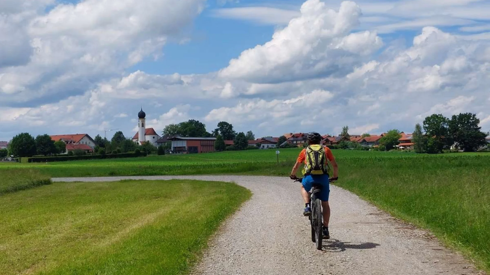 Radfahrer auf Landstraße vor Dorf mit Kirche und blauem Himmel
