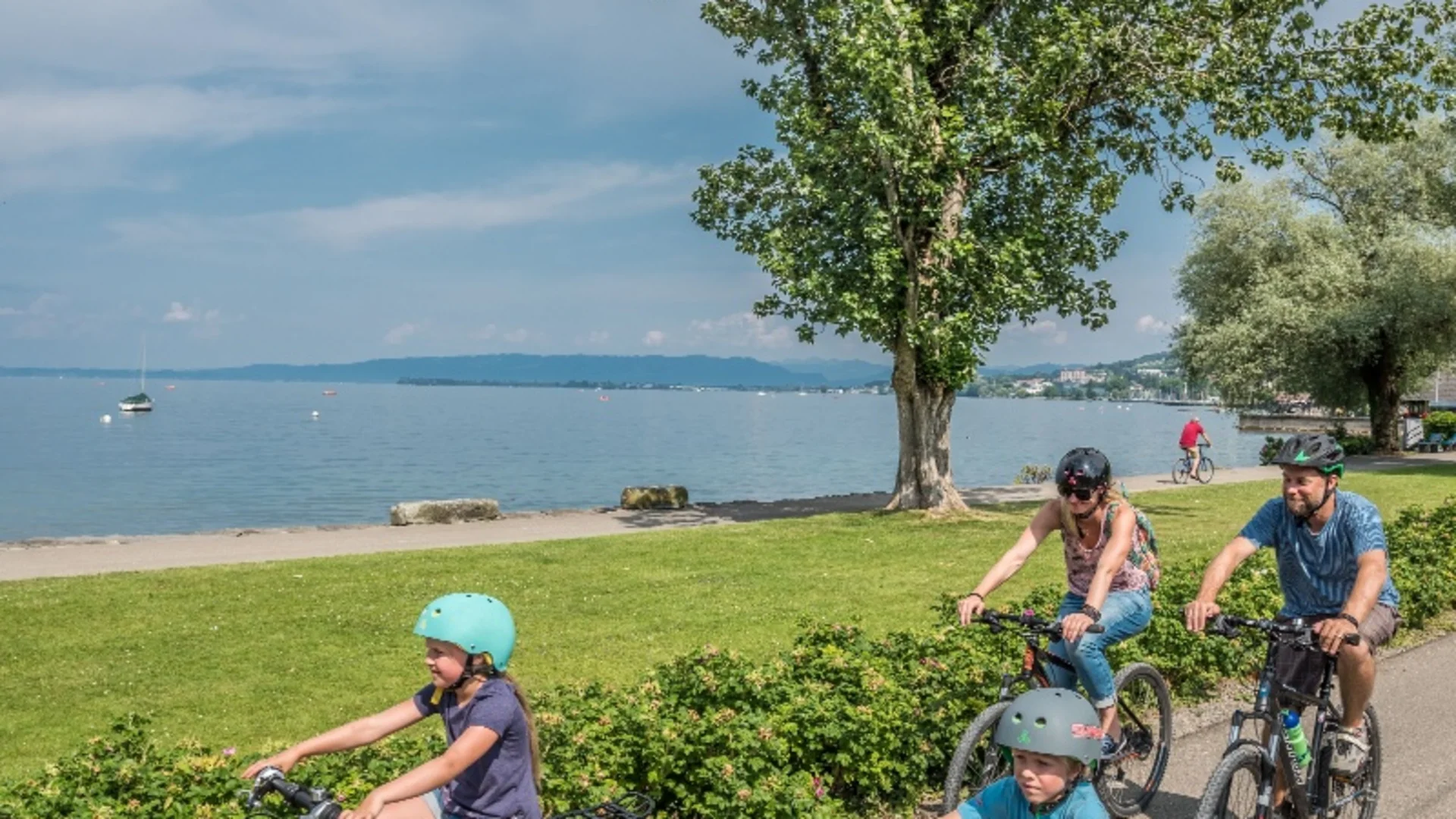 Familie fährt mit Fahrrädern auf einem Weg am Seeufer bei sonnigem Wetter