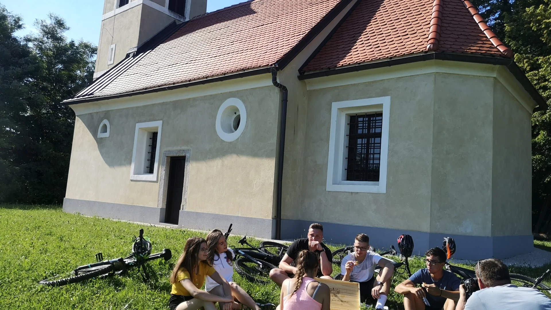 Groep jongeren zittend op gras bij een kleine kerk met fietsen