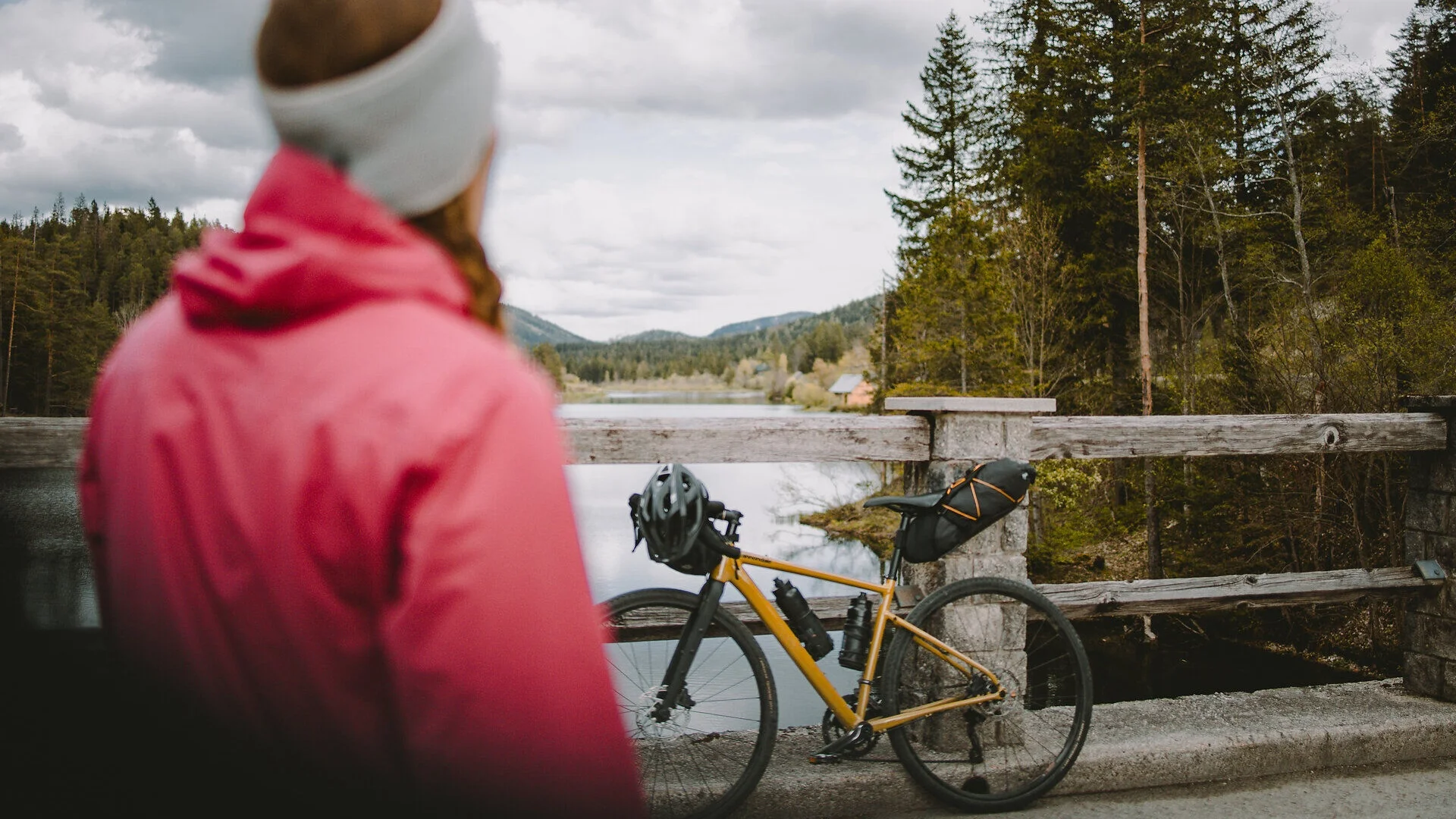 Tour-Detail Person in rotem Mantel blickt auf Fahrrad neben See und Wälder unter bewölktem Himmel