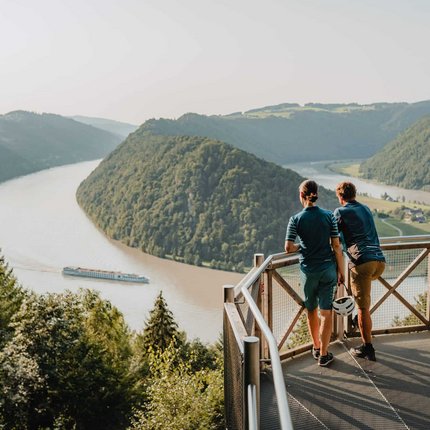 Two people on viewpoint overlooking river, hills, and passing boat