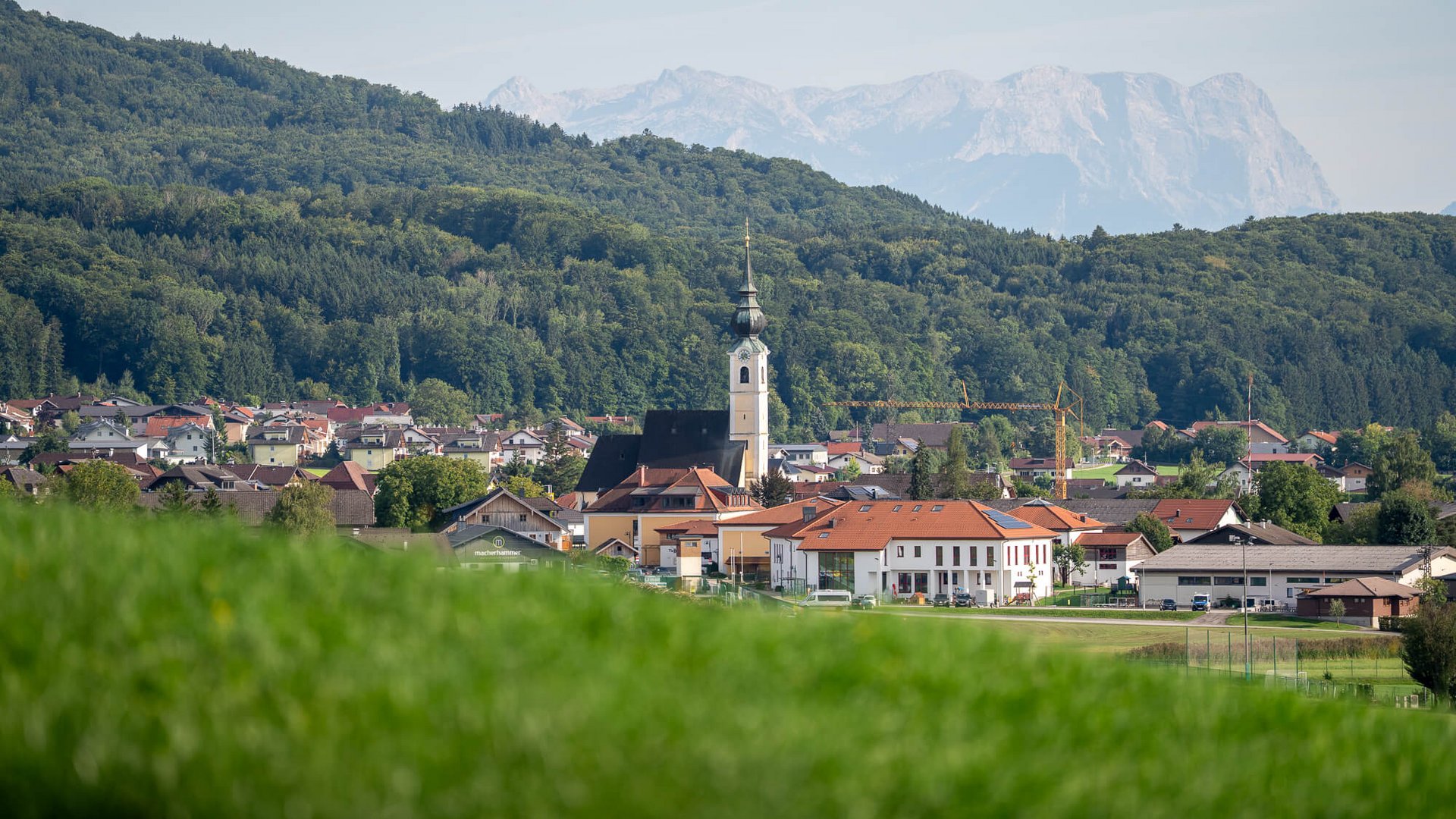 Dorf mit Kirche vor bewaldeten Hügeln und Bergen im Hintergrund