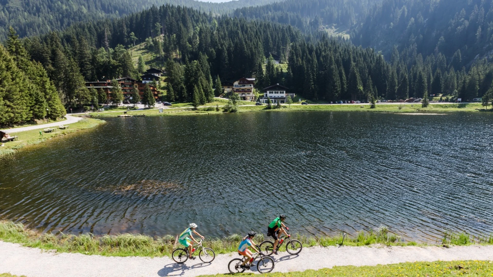 Drei Radfahrer auf einem Weg neben einem See in bewaldeter Berglandschaft