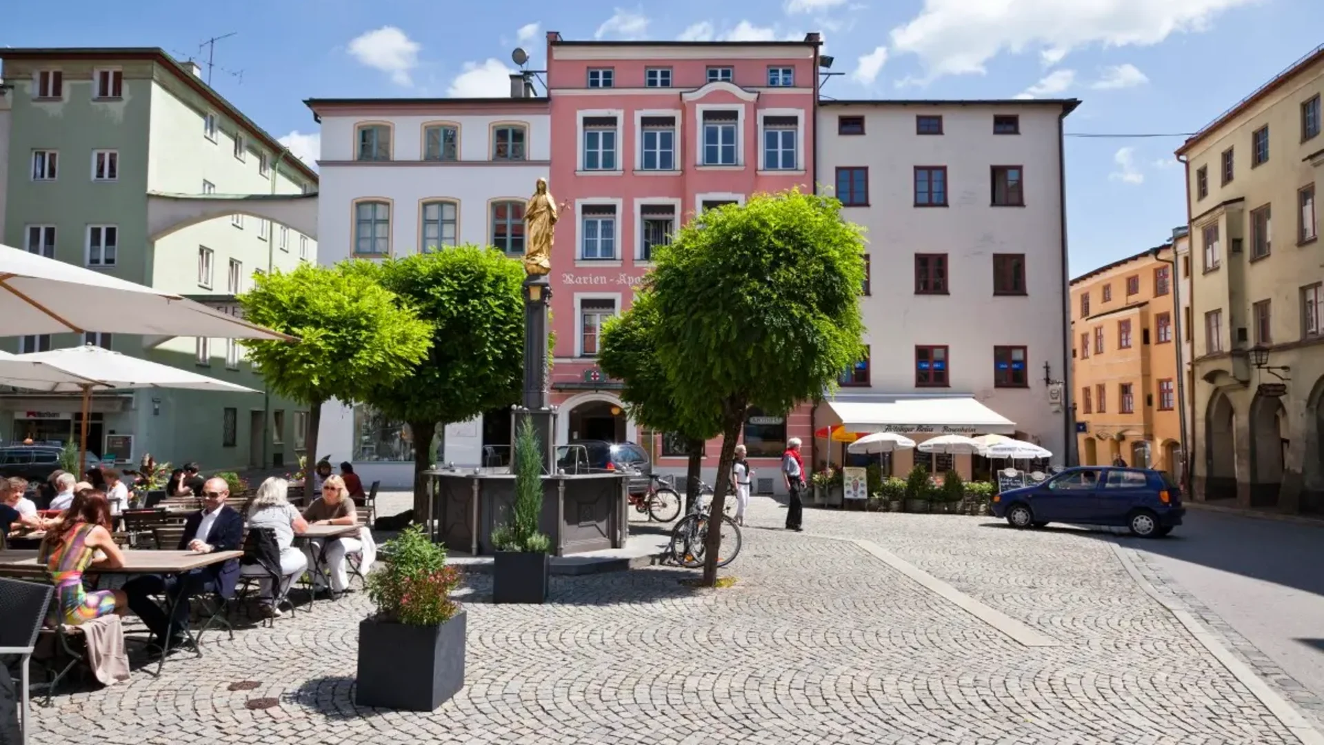 Belebter Platz mit Cafés, Bäumen und bunten Häusern unter blauem Himmel