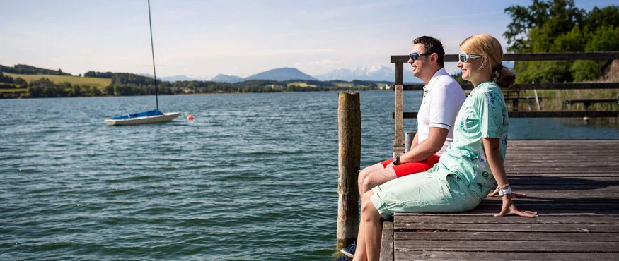 Koppel zittend op een steiger uitkijkend over een meer met zeilboot en bergen