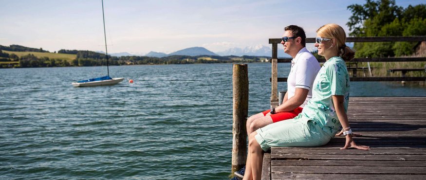Couple sitting on a dock looking at a lake with sailboat and mountains
