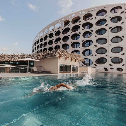 Person swimming in an outdoor pool in front of a modern building with round windows