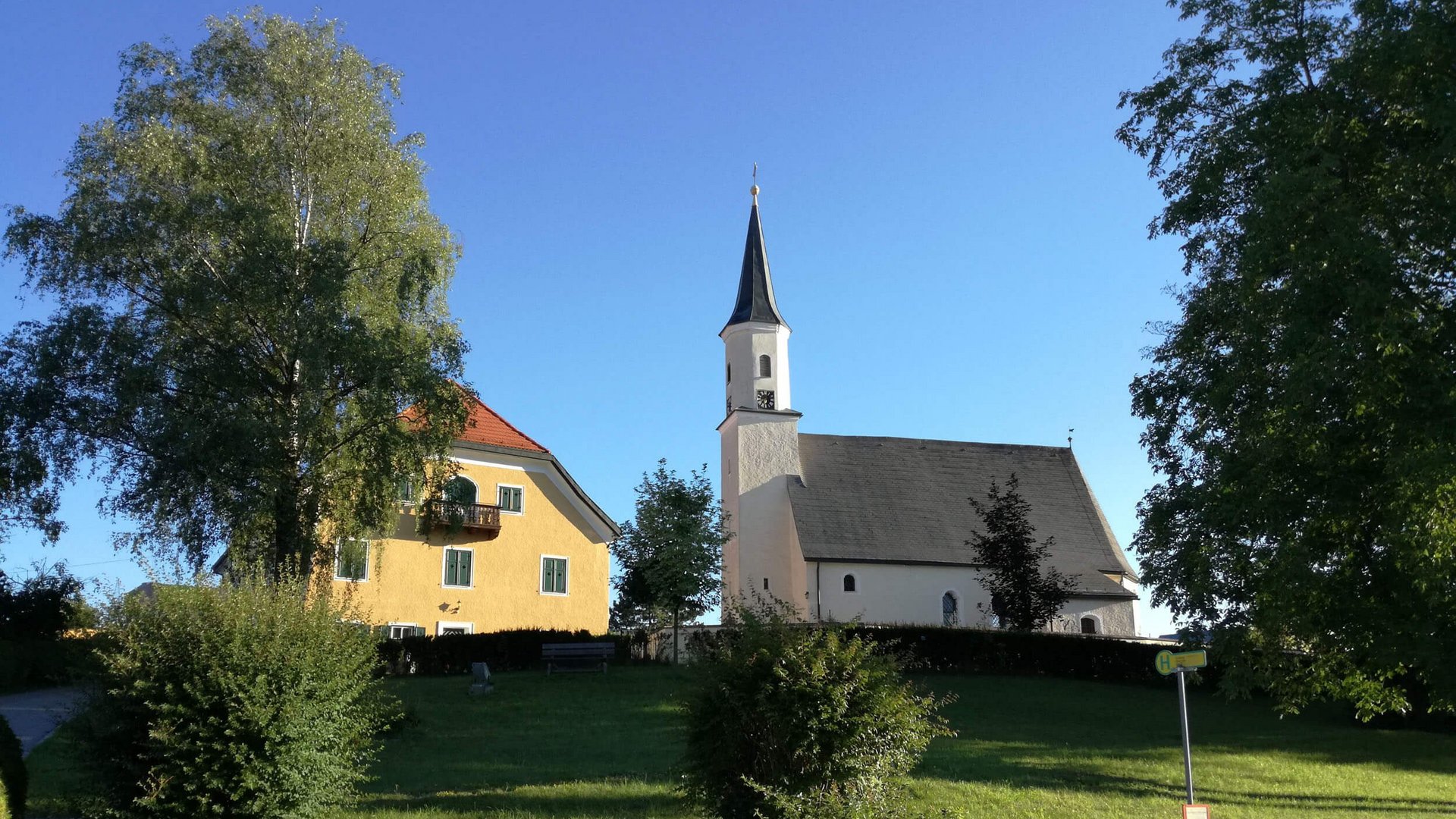 Barocktour © Perwang Kirche Kirche und gelbes Haus auf einem Hügel unter blauem Himmel