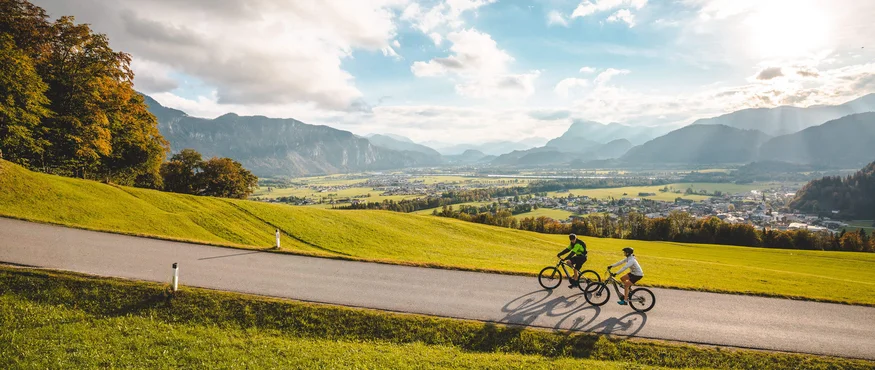 Zwei Radfahrer fahren auf einer Straße durch eine grüne Landschaft mit Bergen