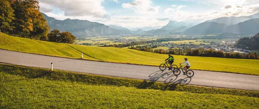 Zwei Radfahrer fahren auf einer Straße durch eine grüne Landschaft mit Bergen