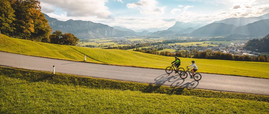 Two cyclists riding on a road through green hills with mountains in the background