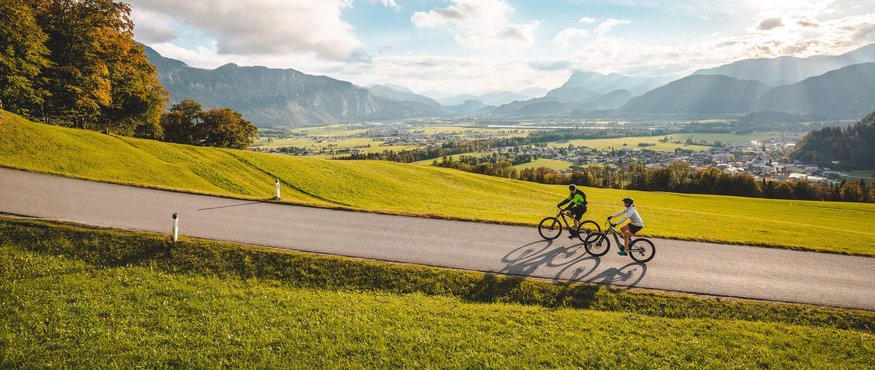 Two cyclists riding on a road through green hills with mountains in the background