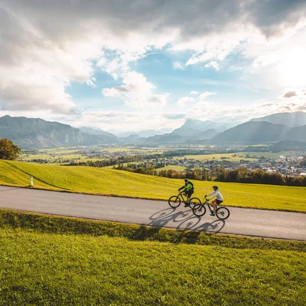 Zwei Radfahrer fahren auf einer Straße durch eine grüne Landschaft mit Bergen