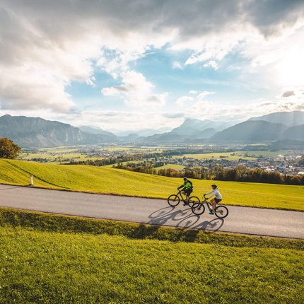 Two cyclists riding on a road through green hills with mountains in the background