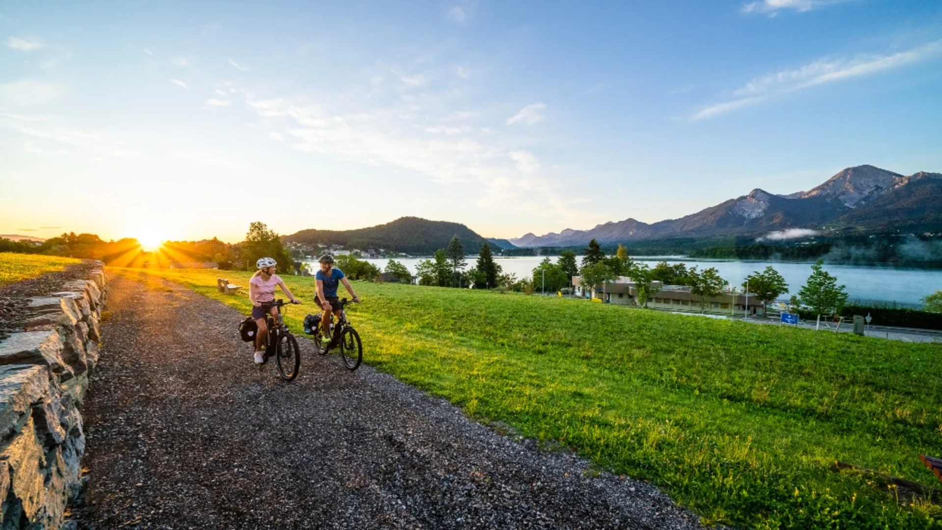 Twee mensen fietsen op een pad bij zonsopgang met uitzicht op bergen en meer
