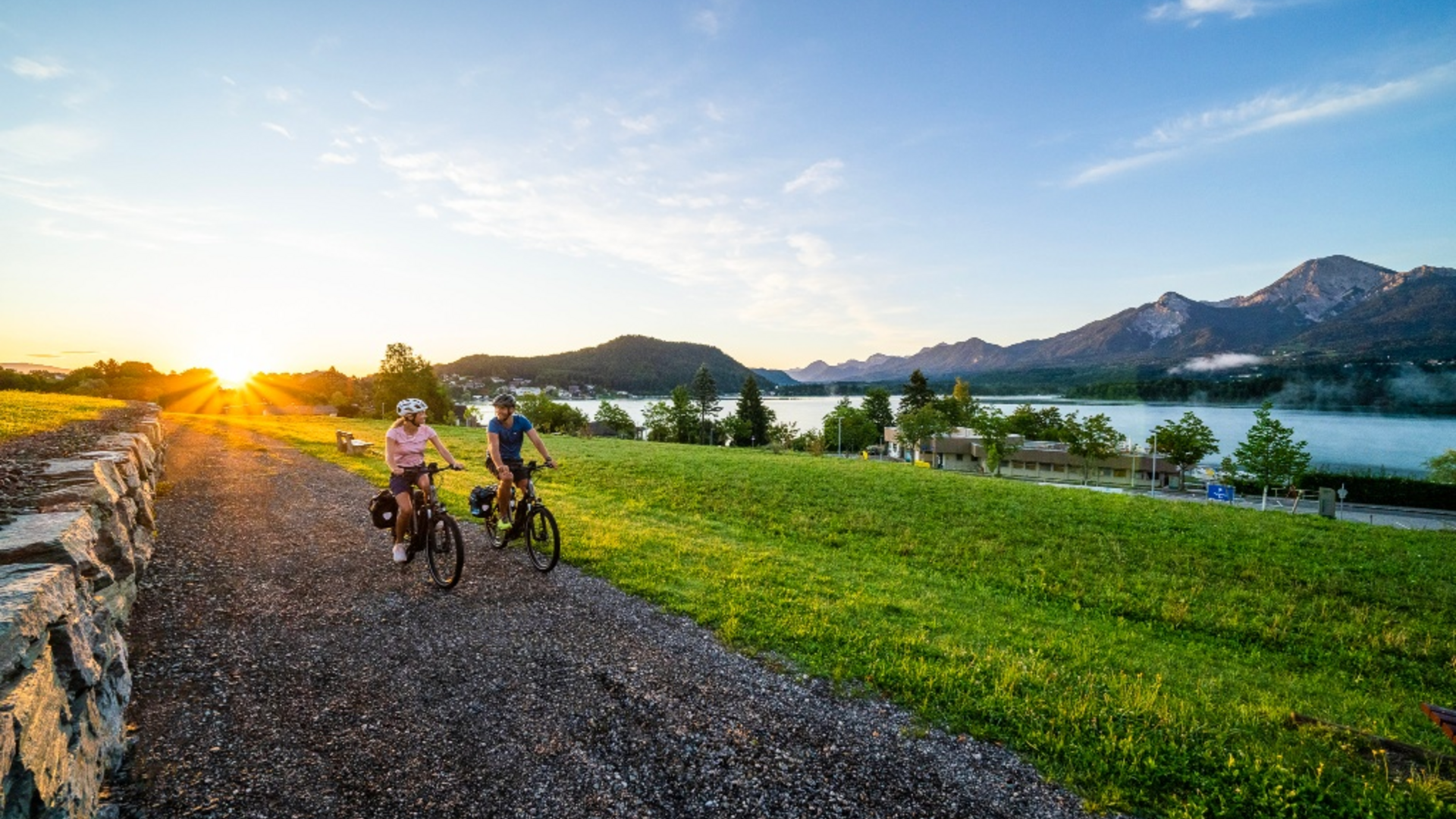 Two people cycling on a path at sunrise with mountain and lake scenery