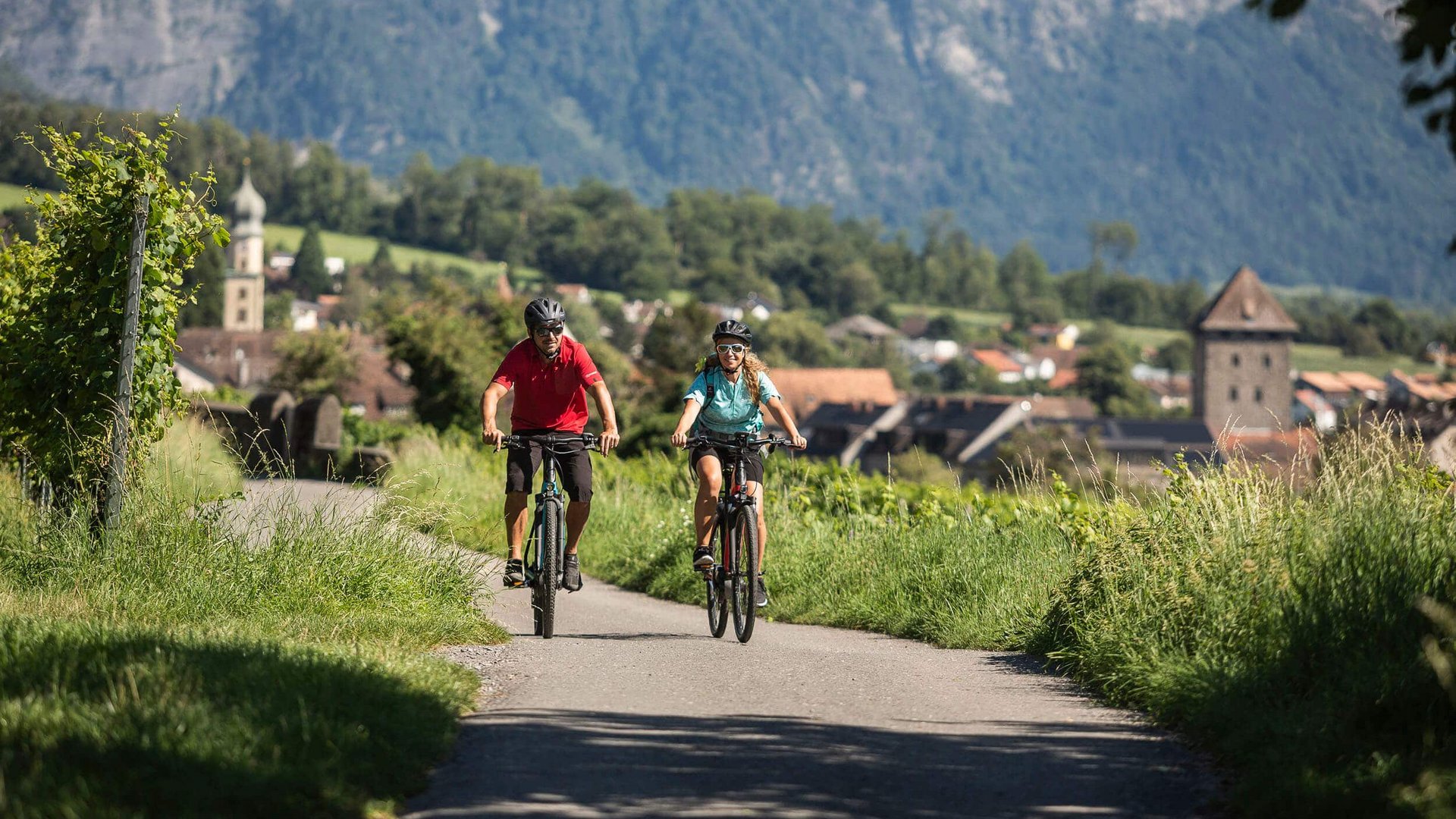 Two cyclists riding on countryside road with mountain village and mountains behind