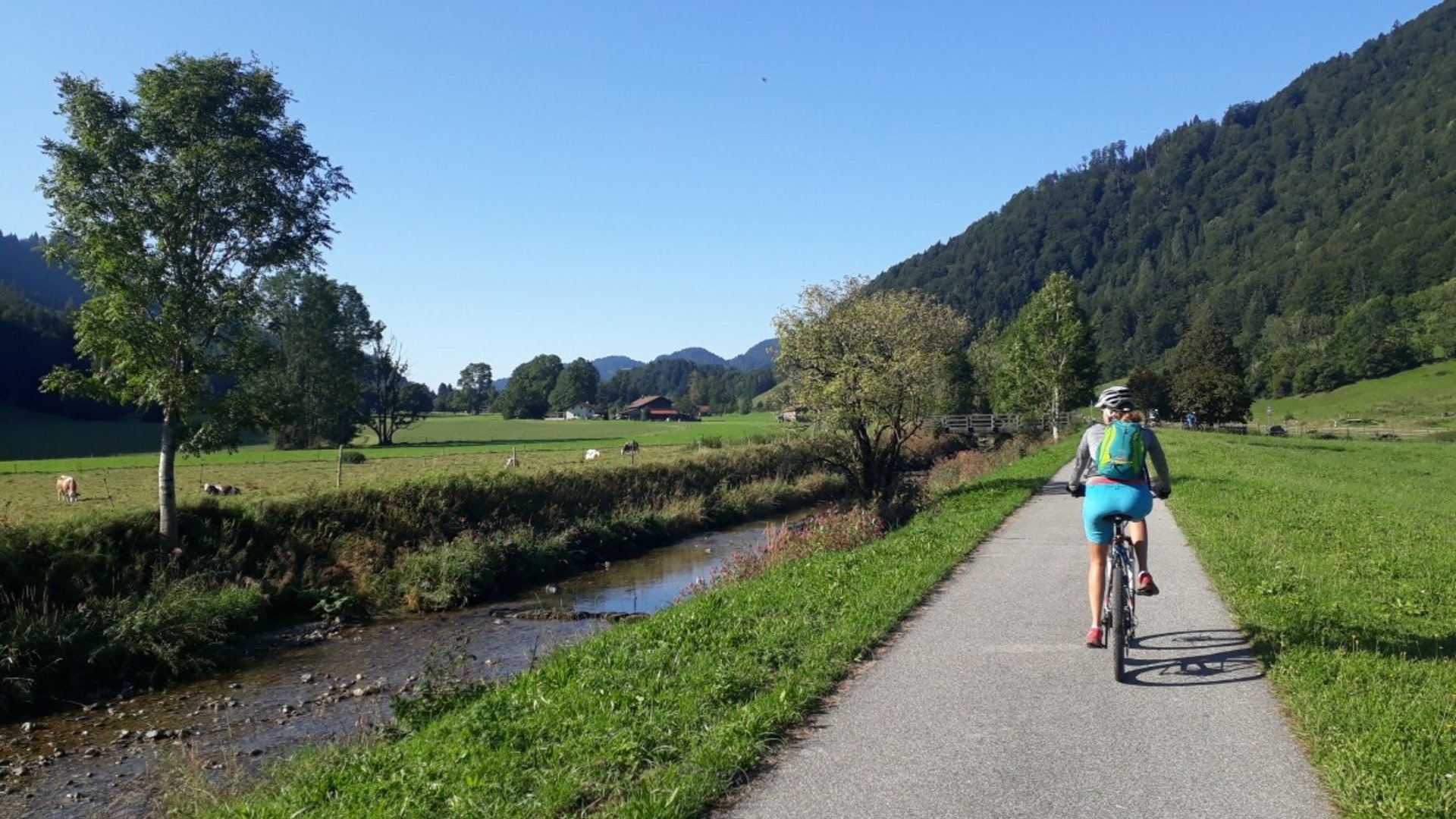 Cyclist riding on path beside stream and fields with mountains in background