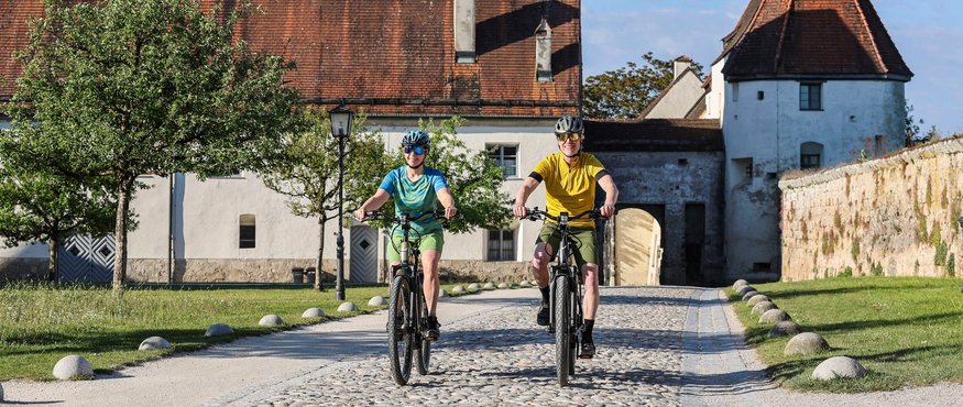 Two cyclists riding on cobblestone path in front of historic building with red tiled roof