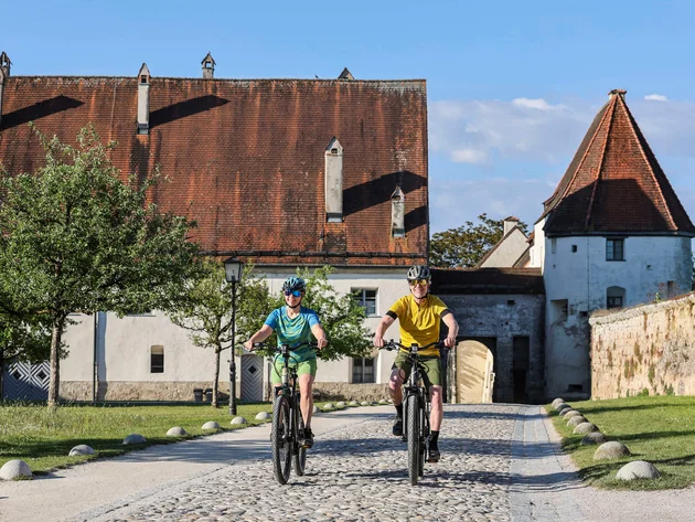 7 Quellen der Entschleunigung © Velontour - Heiko Mandl Zwei Radfahrer vor historischem Gebäude mit rotem Ziegeldach bei Sonnenschein