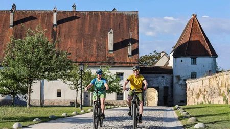 7 Quellen der Entschleunigung © Velontour - Heiko Mandl Zwei Radfahrer vor historischem Gebäude mit rotem Ziegeldach bei Sonnenschein