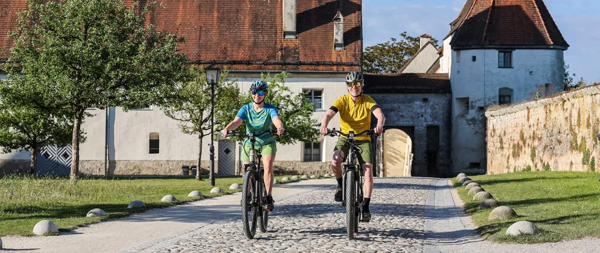 Twee fietsers op kasseienweg voor historisch gebouw met rood dak bij zonlicht