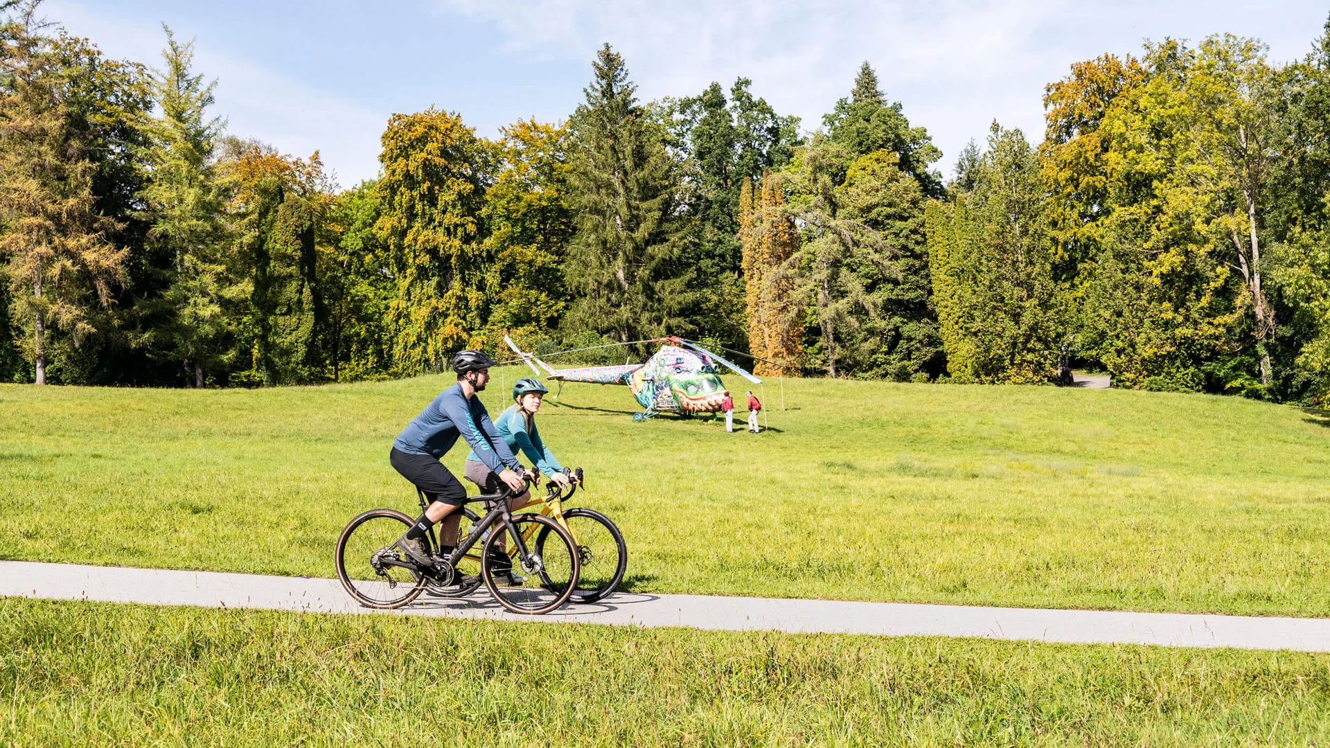 Twee fietsers op een parkpad met een beschilderde helikopter op de achtergrond