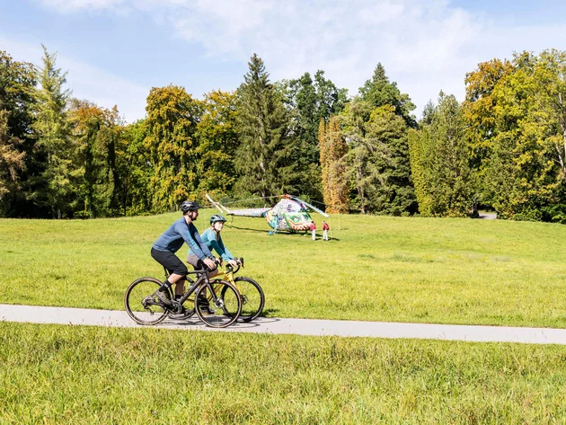 Zwei Radfahrer auf einem Parkweg mit bemaltem Hubschrauber im Hintergrund