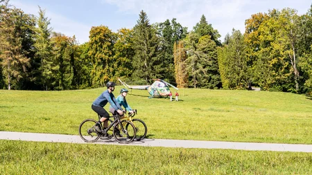 Zwei Radfahrer auf einem Parkweg mit bemaltem Hubschrauber im Hintergrund