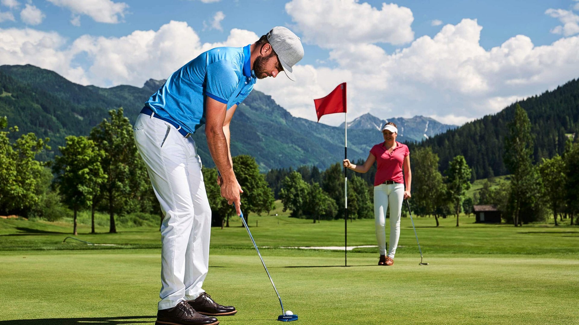 Man playing golf putting while woman holds flag on green golf course with mountains