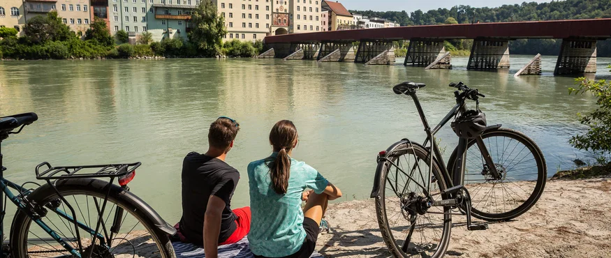 Zwei Personen sitzen am Fluss mit Fahrrädern und Blick auf historische Gebäude