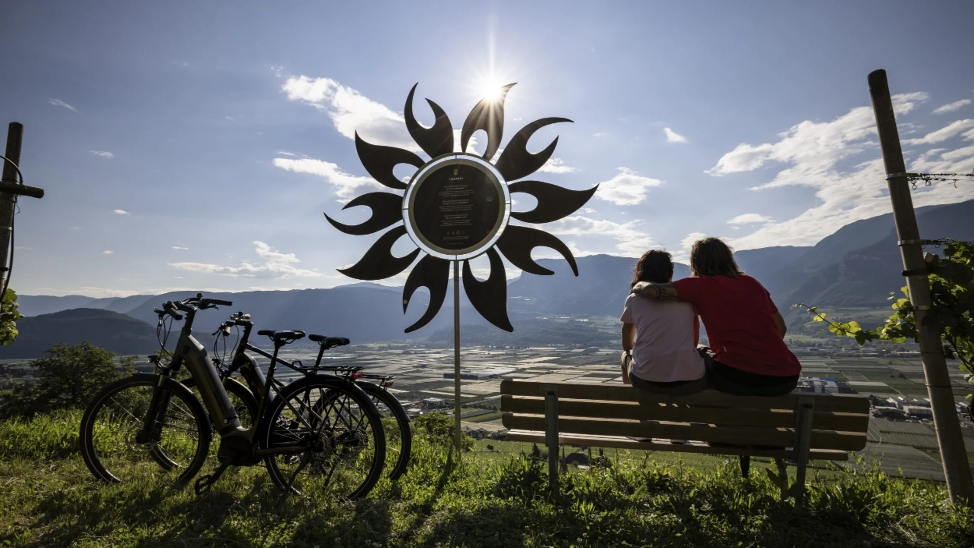 Paar sitzt auf Bank neben Fahrrädern mit Bergtal und Sonnenskulptur im Hintergrund